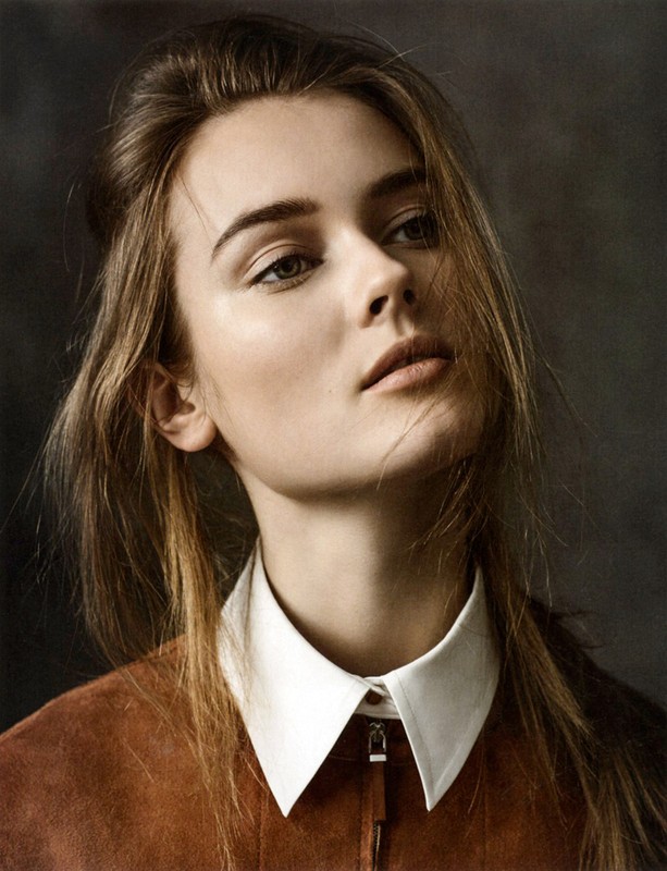 Portrait of a young woman with long brown hair looking upwards, wearing a brown suede jacket and white collared shirt against a dark background.