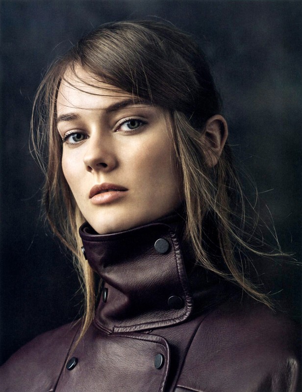 Portrait of a young woman with brown hair wearing a dark plum leather high-neck jacket against a textured dark background.