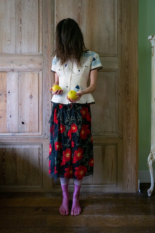 A slender woman stands before a paneled door, face obscured by dark hair, wearing floral skirt, holding yellow apples.