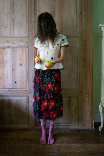 A slender woman stands before a paneled door, face obscured by dark hair, wearing floral skirt, holding yellow apples.