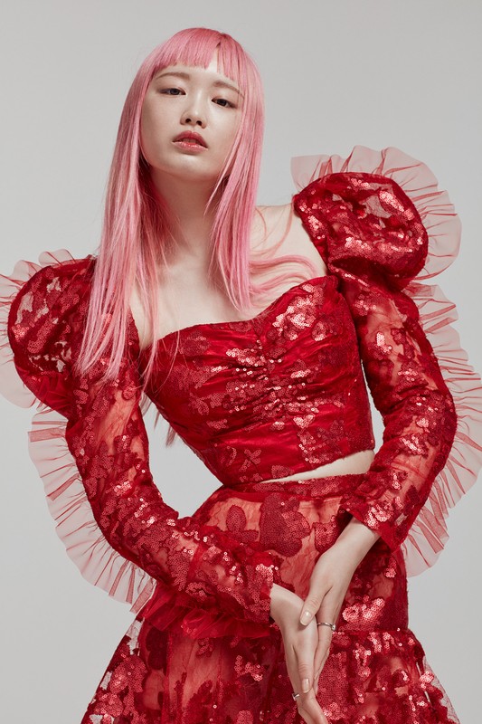 Portrait of an Asian woman with pink hair wearing a red, floral-patterned two-piece outfit against a light gray background.