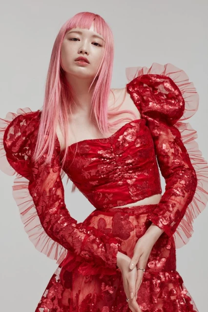 Portrait of an Asian woman with pink hair wearing a red, floral-patterned two-piece outfit against a light gray background.