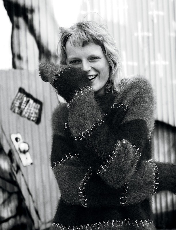 Smiling woman with blonde hair in a black and gray sweater, standing outside in front of a metal wall in black and white.