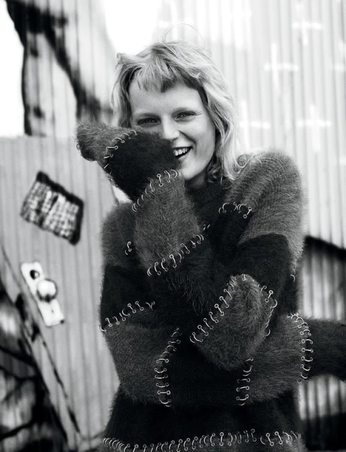 Smiling woman with blonde hair in a black and gray sweater, standing outside in front of a metal wall in black and white.
