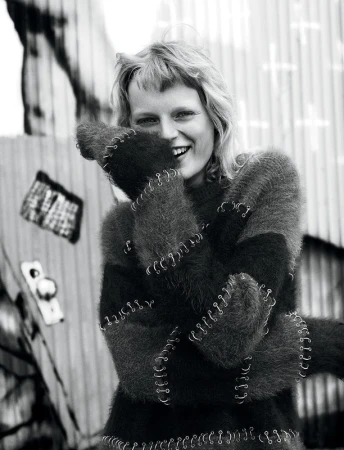 Smiling woman with blonde hair in a black and gray sweater, standing outside in front of a metal wall in black and white.