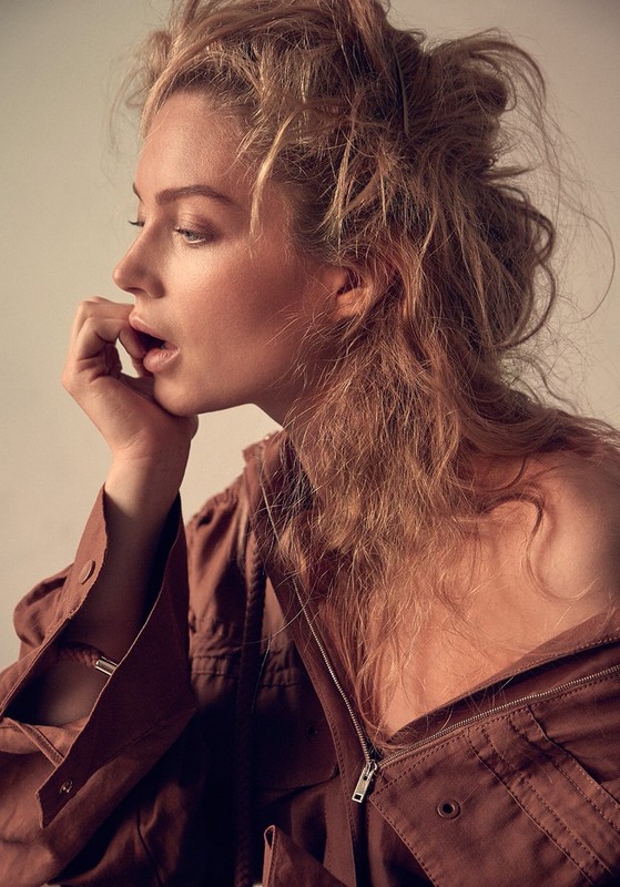 Blonde woman in unzipped brown jacket with hand at face poses in a medium shot indoors with neutral lighting.