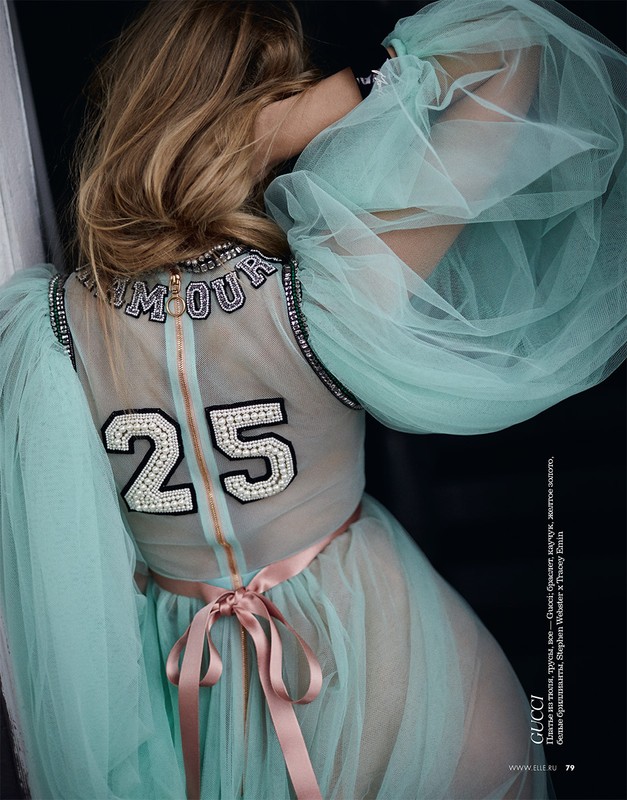 Blonde fashion model wearing a sheer aqua dress with embellishments posing in a studio. Soft diffused light accentuates the fabric.