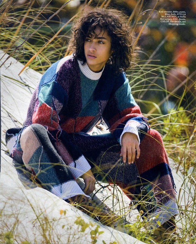 An adult female poses in an outdoor grassy field wearing a multi-colored patchwork sweater and matching pants, creating an editorial fashion shot.