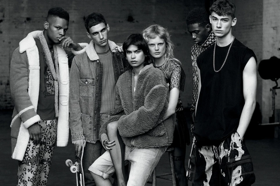 Group of six fashion models in monochromatic studio shot showcasing diverse outerwear and casual streetwear against a neutral backdrop.