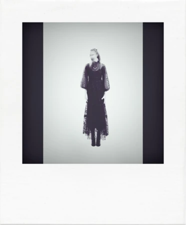 Monochromatic fashion shot featuring a woman in a long, dark, lace dress against a pale background, framed by a black vertical bar.