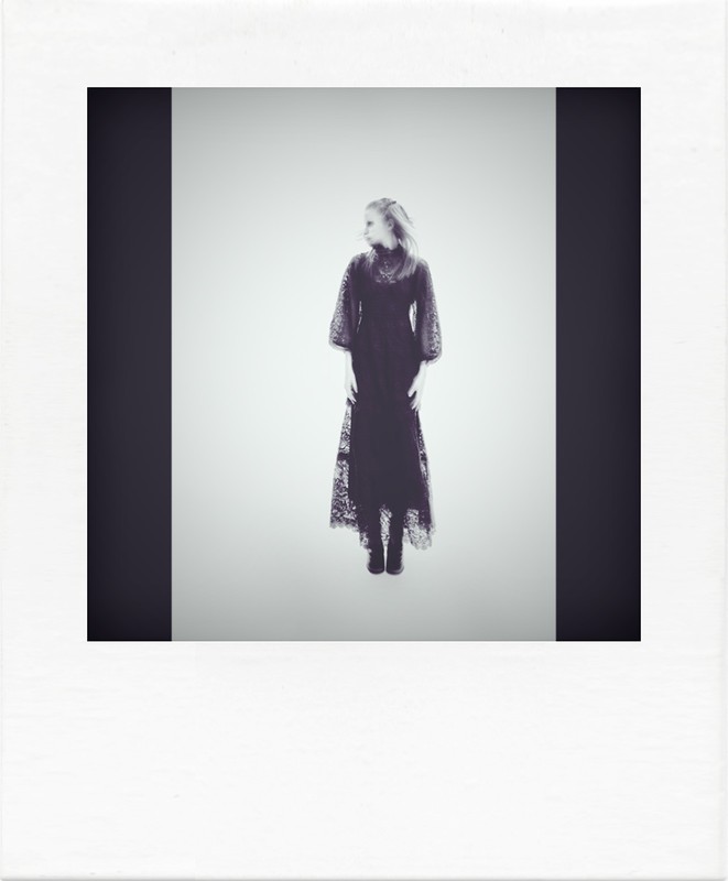 A single woman in a long black dress stands in a studio looking over her shoulder; presented inside of a white instant-film frame.