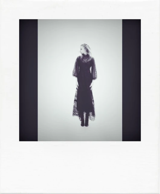 A single woman in a long black dress stands in a studio looking over her shoulder; presented inside of a white instant-film frame.