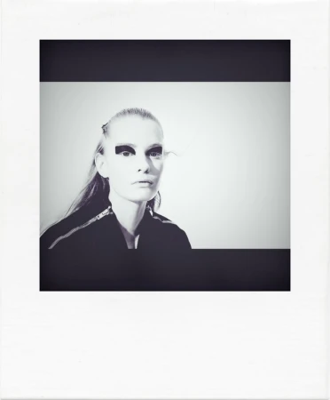 Black and white portrait of a model with dark eye makeup and slicked hair inside a polaroid frame.