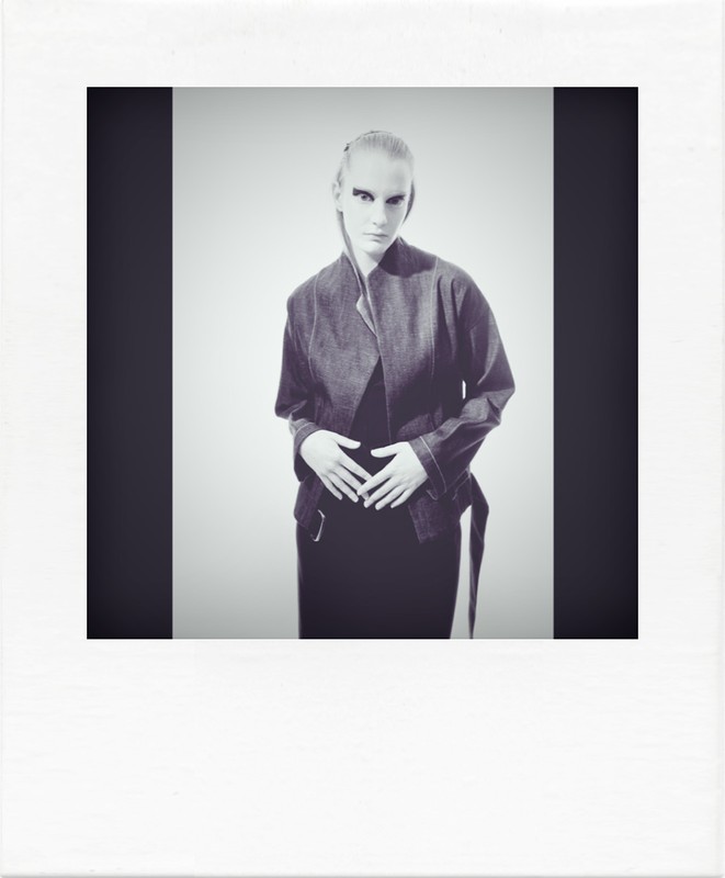 Studio portrait of a woman with pale skin wearing a jacket, standing against a gray background, framed by dark vertical bands.