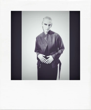 Studio portrait of a woman with pale skin wearing a jacket, standing against a gray background, framed by dark vertical bands.