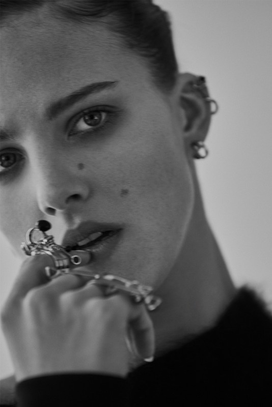 Black and white portrait of a young woman with short hair and multiple ear piercings wearing statement rings while looking at the camera.