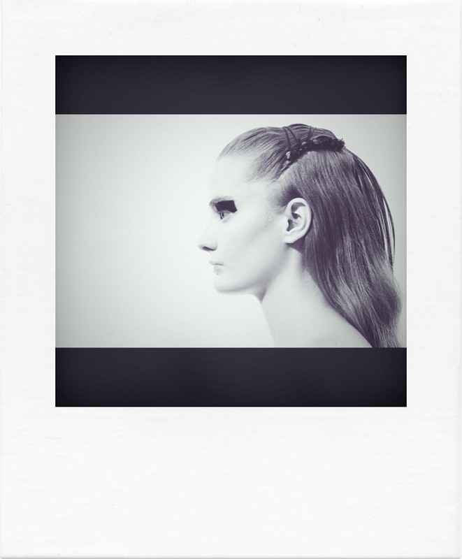 Portrait of a fair-skinned adult female with dark eye makeup and styled hair, photographed in profile against a light background.
