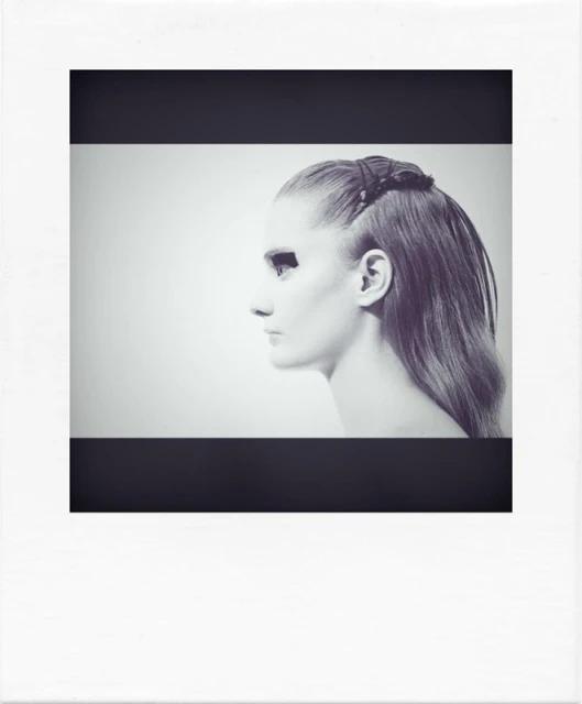 Portrait of a fair-skinned adult female with dark eye makeup and styled hair, photographed in profile against a light background.