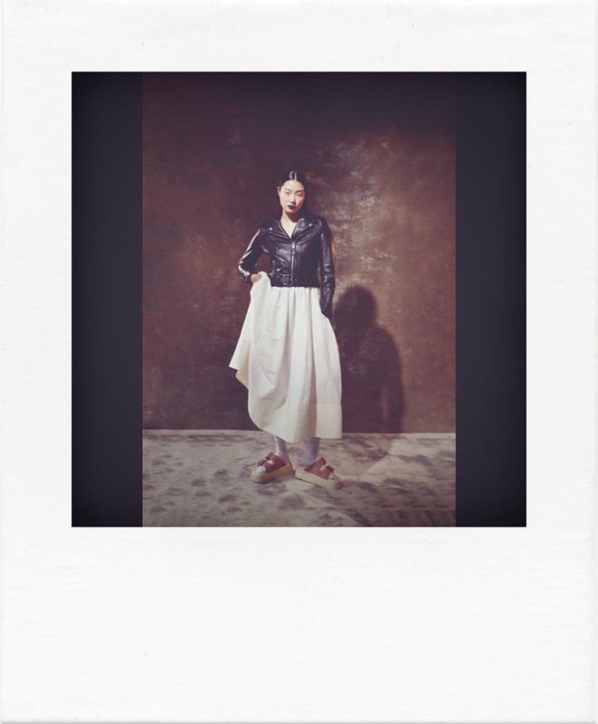 Polaroid photograph of a model in a black leather jacket and white skirt standing against a textured backdrop