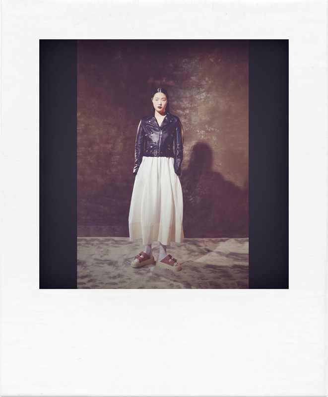 Full shot of an adult woman in a black leather jacket and white dress, posing in a photo studio with a textured backdrop and soft lighting.