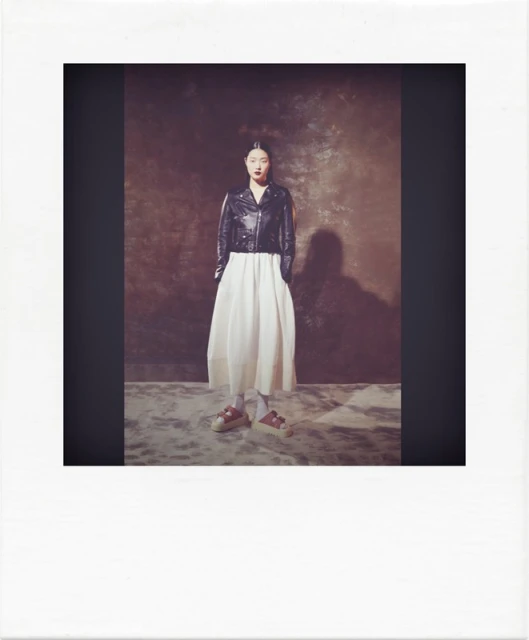Full shot of an adult woman in a black leather jacket and white dress, posing in a photo studio with a textured backdrop and soft lighting.