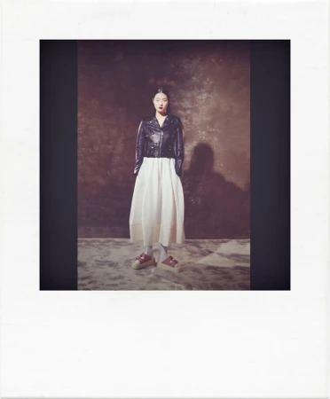 Full shot of an adult woman in a black leather jacket and white dress, posing in a photo studio with a textured backdrop and soft lighting.