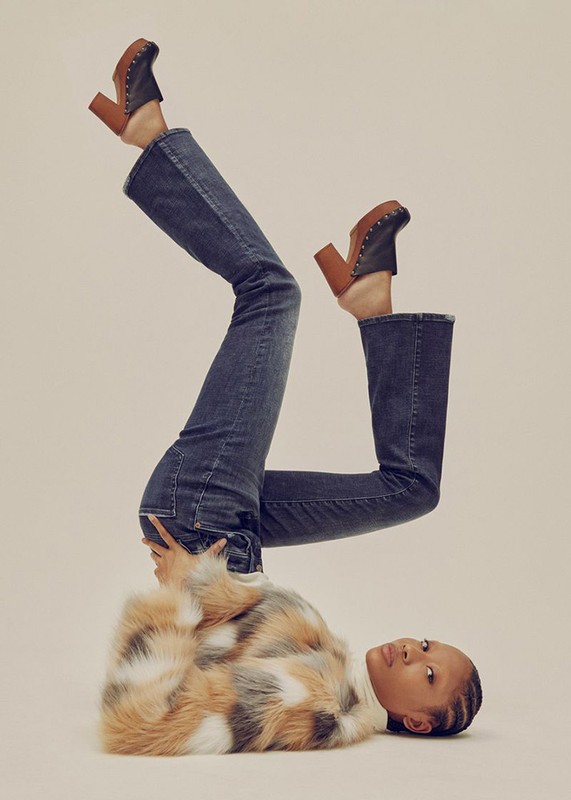 Woman in a fur coat, blue jeans, and brown clogs poses upside down against a light background in a fashion editorial shot.