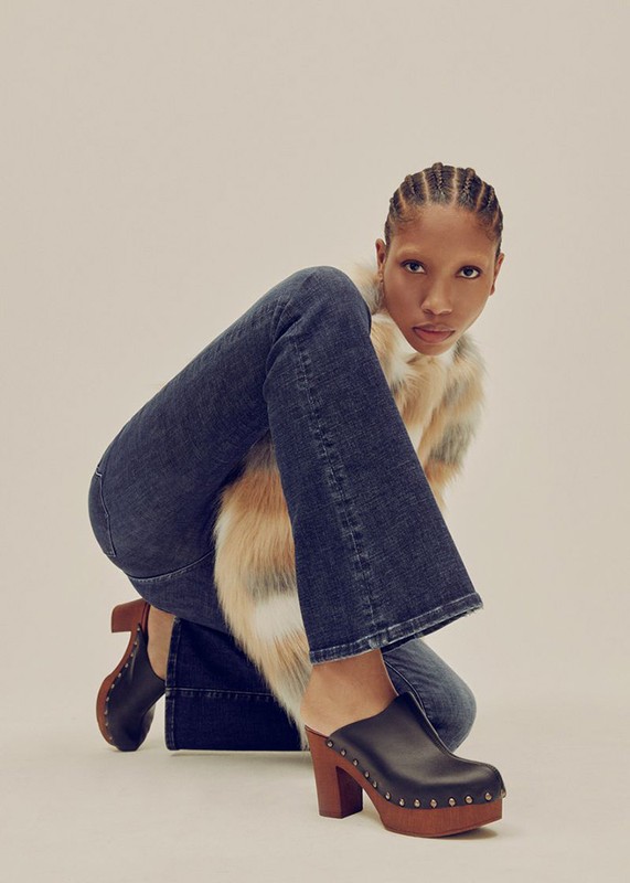 Woman poses in a furry vest, blue jeans, and brown platform shoes in a studio setting with soft lighting, showcasing a retro fashion style.