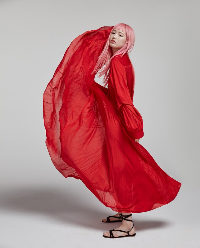 Elegant Asian woman with pink hair in a red dress poses in studio with flowing fabric, looking at camera. Fashion editorial shot.