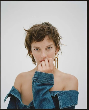 Portrait of a woman with short tousled brown hair wearing an off-the-shoulder denim jacket against a plain white background.