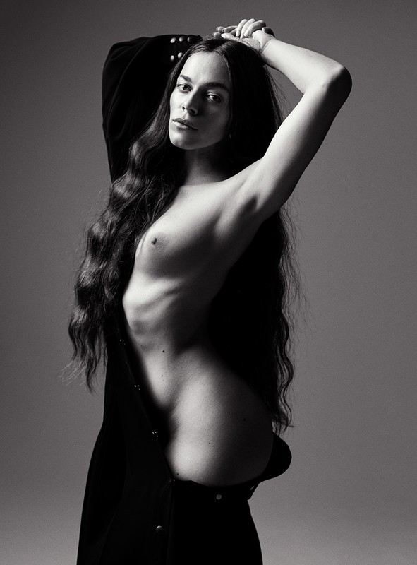 Black-and-white portrait of a slender woman with long wavy hair, partially unclothed, posing against a neutral background, creating a dramatic effect.