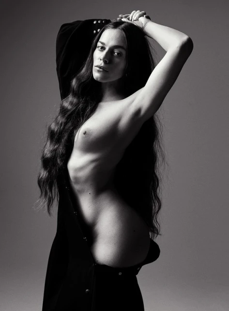 Black-and-white portrait of a slender woman with long wavy hair, partially unclothed, posing against a neutral background, creating a dramatic effect.