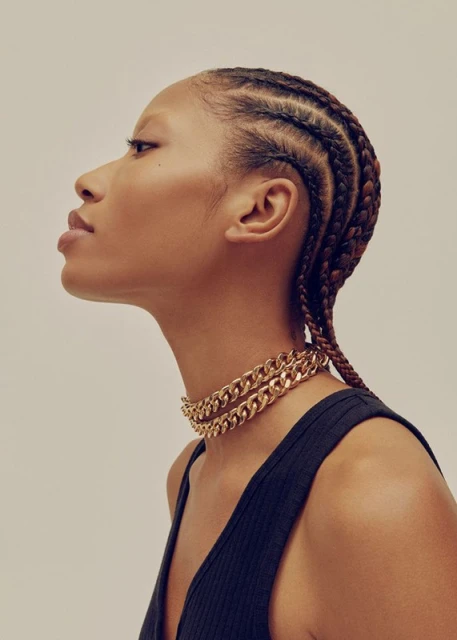 Profile shot of a woman with braided hair wearing a gold chain necklace against a light background in a beauty or fashion context.