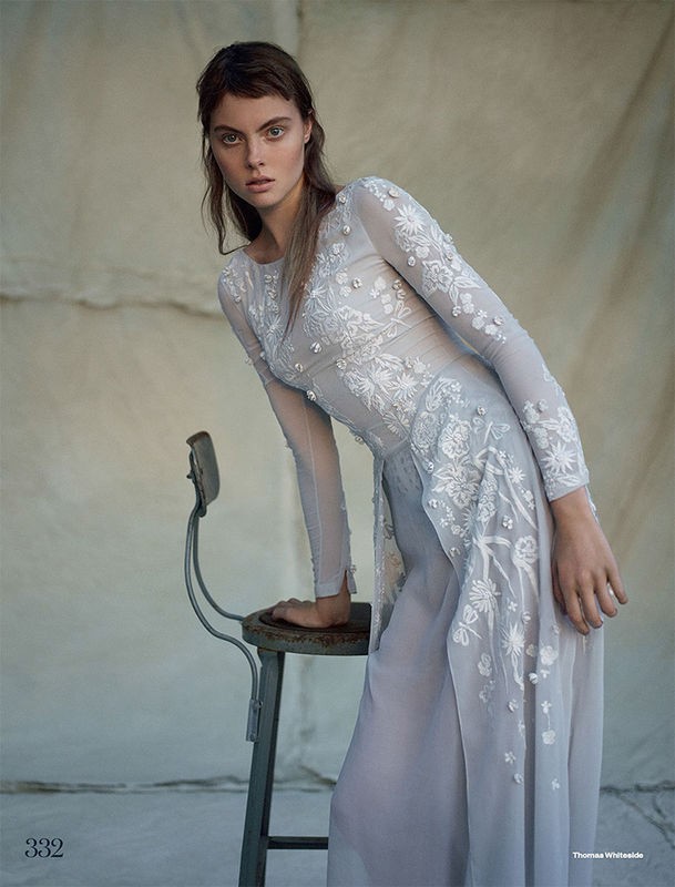 Fashion editorial shot of a female model in a light blue, floral-embroidered gown, posed next to an old stool in a muted setting.