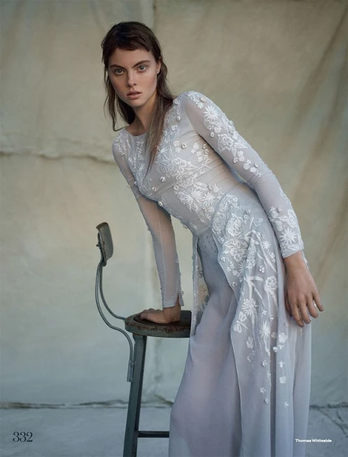 Fashion editorial shot of a female model in a light blue, floral-embroidered gown, posed next to an old stool in a muted setting.