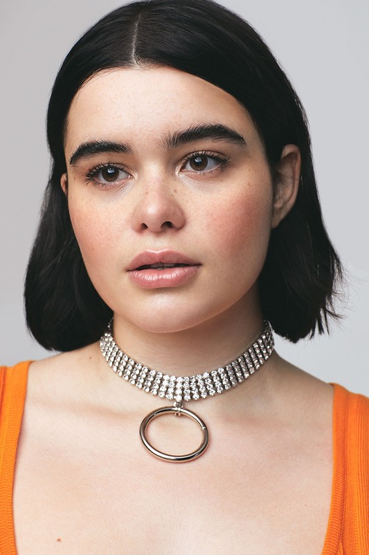 Portrait of a young woman with dark hair wearing a rhinestone choker necklace and an orange top against a plain light background