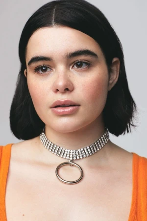 Portrait of a young woman with dark hair wearing a rhinestone choker necklace and an orange top against a plain light background
