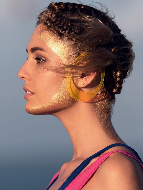 Profile portrait of a woman with braided hair and gold body paint wearing transparent yellow hoop earrings against a blurry background.