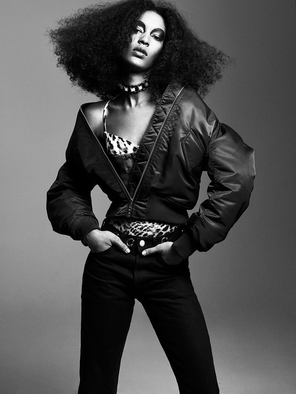 Black and white studio portrait of an adult female fashion model with afro hairstyle wearing an animal print top and bomber jacket.