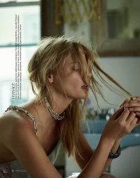 Close-up portrait of a blonde woman with messy hair styling, wearing a silver chain choker necklace and dress in an indoor setting.