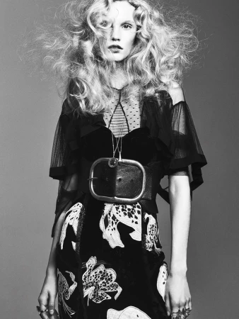 Model with voluminous blonde hair wearing a black dress and oversized buckle belt against a grey background