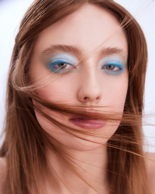 Close-up portrait of a woman with blue eye shadow and neutral lipstick, with hair partially covering her face, against a white background.