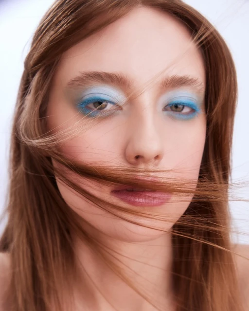 Close-up portrait of a woman with blue eye shadow and neutral lipstick, with hair partially covering her face, against a white background.