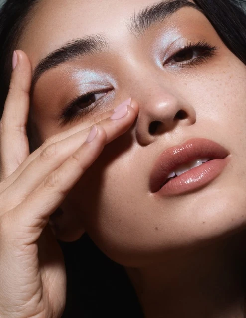 Close-up portrait of an Asian adult female model with light blue and tan eyeshadow, a nude lip, and glowing skin holding her hand to her face.