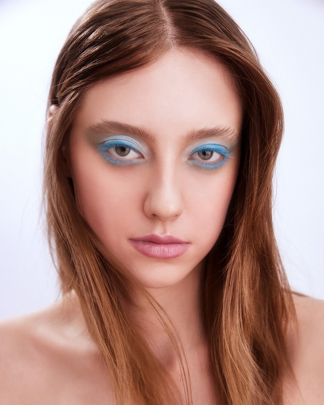 Close-up portrait of a young woman with light brown hair and blue eyeshadow on a light backdrop for beauty and fashion concepts.