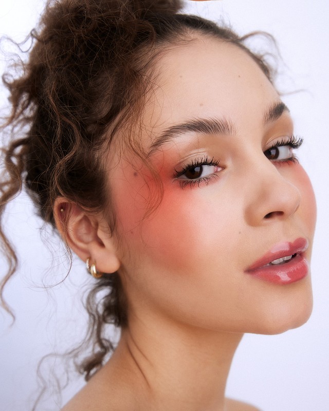 Close-up portrait of a young adult female with curly hair and vibrant red blush, against a clean white backdrop in a beauty shot.