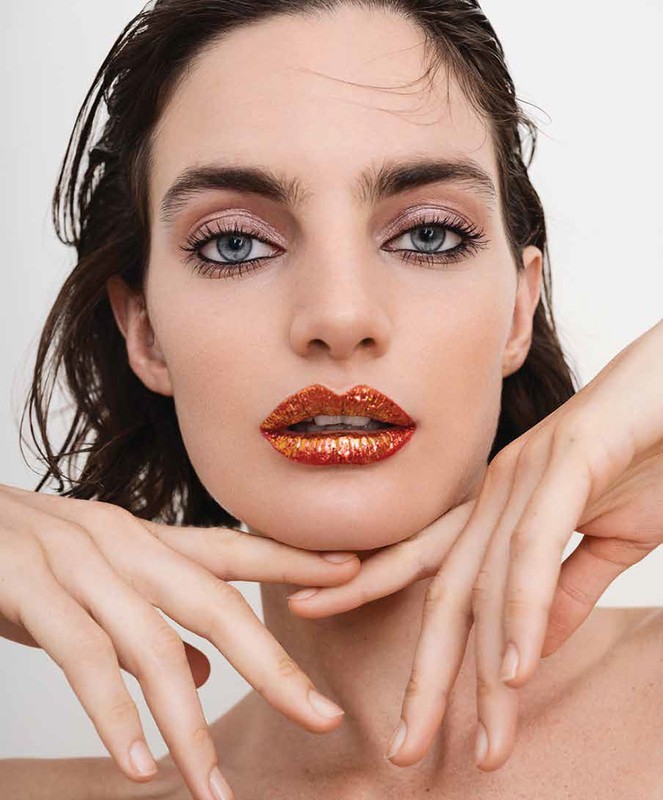 Close-up beauty shot of a woman with sparkling orange lipstick, hands near her face, soft lighting, set against a white backdrop.