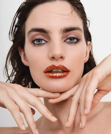 Close-up beauty shot of a woman with sparkling orange lipstick, hands near her face, soft lighting, set against a white backdrop.