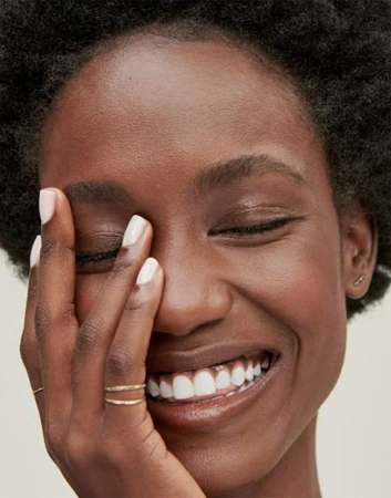 Close-up portrait of a smiling Black woman with short afro hair touching her face with manicured white nails.