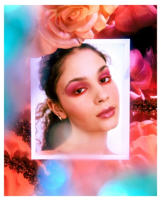 Close-up portrait of a young woman with pink eye shadow and glossy lips, surrounded by soft-focus floral elements.